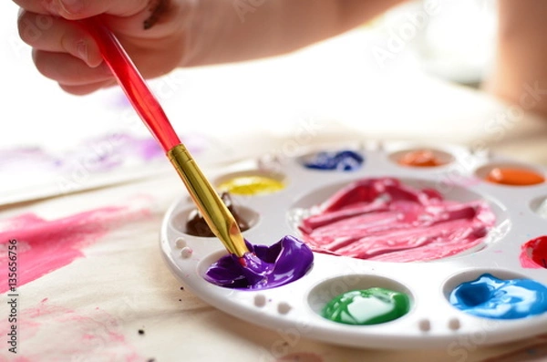 Obraz Child's hand holding a paintbrush dipping into a palette full of colorful paint