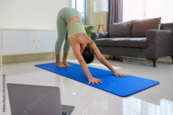 Fototapeta Woman in green activewear performing downward facing dog yoga pose on blue mat in living room with laptop in foreground. Asian female stretching  shoulders chest and hamstrings in morning at home