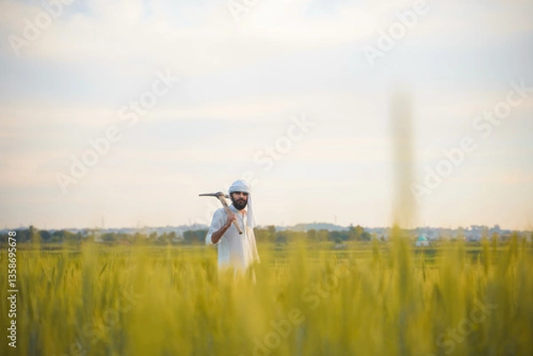 Fototapeta A farmer in traditional white clothing stands in a lush wheat field, holding a pickaxe on his shoulder with a distant village in the background.
