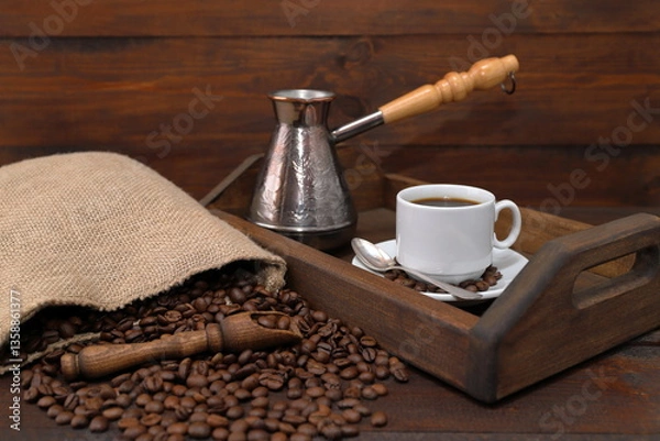 Fototapeta On a brown wooden background there is a wooden tray with a white cup of coffee and a copper turk, next to a scattering of coffee beans from a burlap sack and a wooden scoop with coffee beans. 