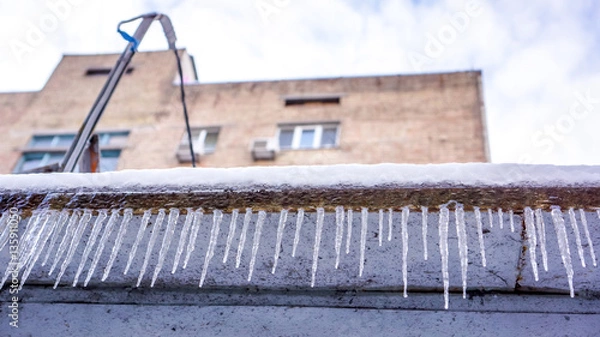 Obraz Winter icicles as fence against multi-storey house and sky with white clouds. Winter hanging icicles on the house roof