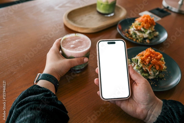 Obraz Person holding a smartphone with blank white screen and a colorful layered drink in a cafe, with plates of gourmet food and cozy interior in the background