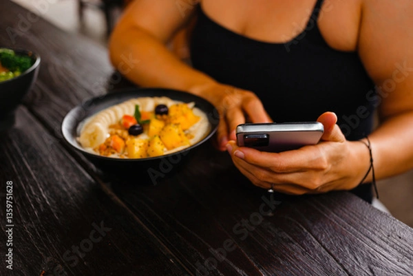 Fototapeta A woman has breakfast of porridge with fruits, berries and holds a phone in her hands. The concept of counting calories using a mobile application.