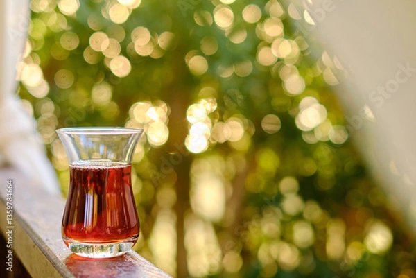 Fototapeta Glass of tea resting on wooden balcony, sunlight filtering through lush green leaves, creating warm and peaceful atmosphere of relaxation and serenity