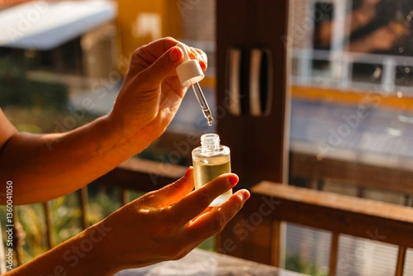 Fototapeta Hand holds a pipette with natural care oil for the face and hair against the background of the morning sunlight. Woman's hand opens a bottle of moisturizing serum based on oils