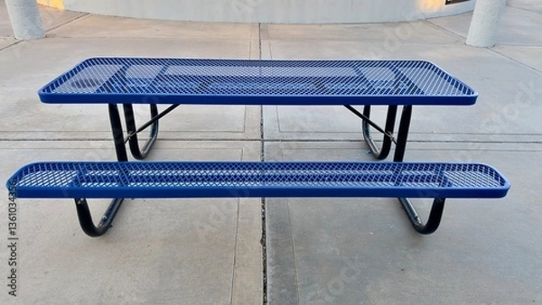Fototapeta empty metal picnic table sits near closed concession stand in quiet sports park shadows stretch across concrete ground breeze moves litter nearby silent pause between games and gatherings