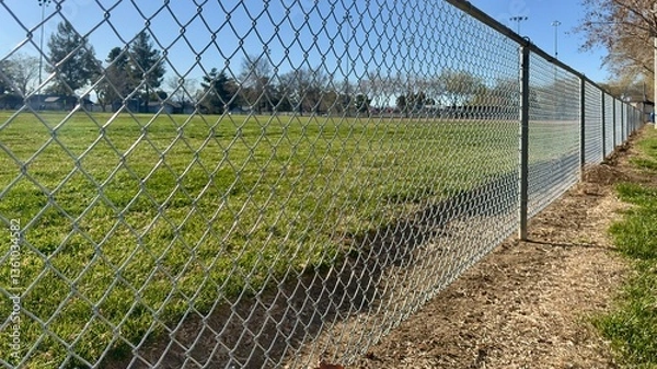 Fototapeta empty sports park enclosed by chain link fence under cloudy sky metal posts stretch along gravel path silent field beyond holds echoes of games past stillness rests on worn bleachers and grass