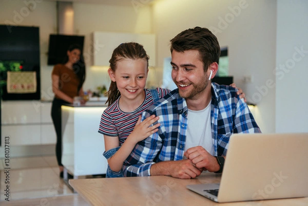 Fototapeta Work from home. Daughter interrupting her father while he is having a business online conversation on his laptop while sitting in the modern living room.