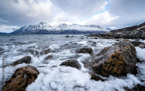 Fototapeta Waves crash against snow-covered rocks near a mountain range under a cloudy sky. The water is rough, and the scene is cold and dramatic