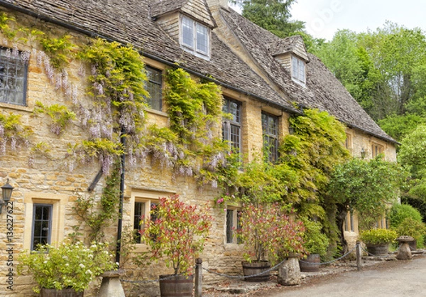 Obraz Charming Cotswolds golden cottages with climbing purple wisteria, plants in the barrels, ornamental stone mushrooms