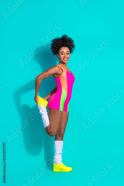 Fototapeta Stylish young woman wearing colorful activewear stretching against vibrant teal vertical background