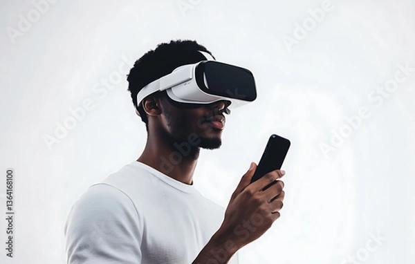 Fototapeta A man wearing VR goggles, holding his phone as if controlling the experience on a white backdrop 