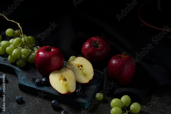 Fototapeta Close-up of a halved apple, red apples, green grapes, and blueberries on a chopping board on a black ceramic table