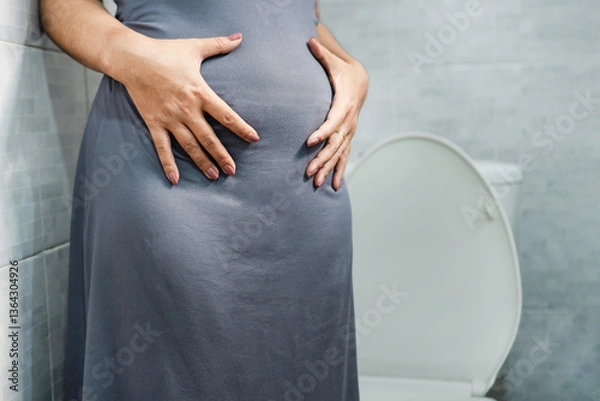 Obraz woman suffering from chronic constipation and fecal impaction, holding her bloated stomach while standing in front of a toilet, feeling discomfort and difficulty due to digestive system problems