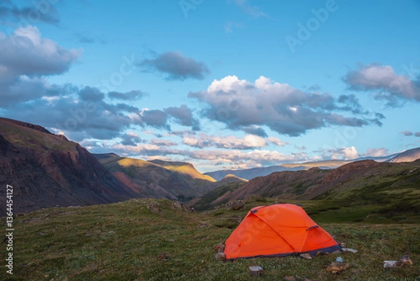 Fototapeta Vivid orange tent on grassy hill with view to mountain tops illuminated by sunset light. Rocks silhouettes. Shadows of clouds on rocky ridge in sunrise colors. Red tent and sunset tones in blue sky.