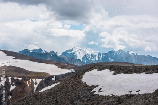 Fototapeta Awesome aerial view to sunlit big stony ridge with snowfield and sheer crags against snow-covered mountain range with snow-capped peaked top. High rocky pass and large snowy pointy peak in cloudy sky.