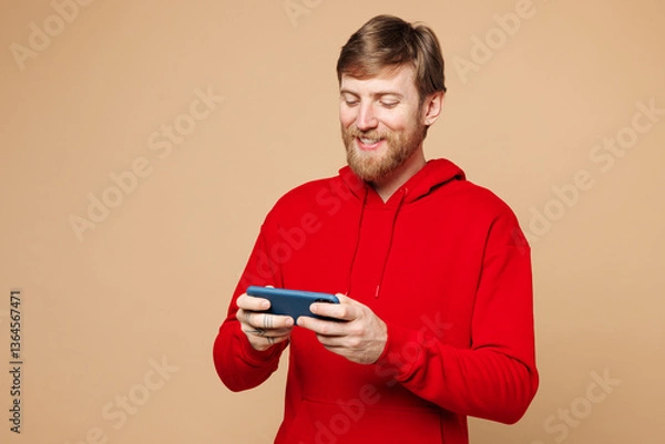 Fototapeta Young man he wear red hoodie casual clothes use play racing app on mobile cell phone hold gadget smartphone for pc video games isolated on pastel plain light beige background studio Lifestyle concept