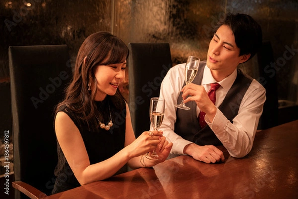 Fototapeta A man and woman dressed formally are smiling at each other while toasting with champagne glasses. They are seated across from each other at a wooden bar with warm ambient lighting.