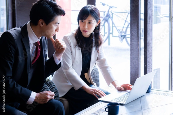 Fototapeta A businessman and a businesswoman in formal attire have a serious conversation while seated by the window at a modern café. Natural light highlights their focused expressions as they engage in a profe