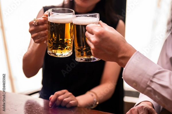Fototapeta A close-up shot of a man and woman toasting with beer mugs in a warmly lit bar. The image focuses on their hands and glasses, emphasizing the golden color of the beer and the festive atmosphere.