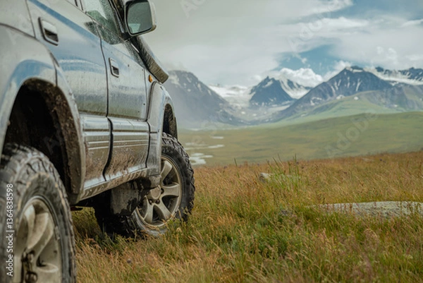 Fototapeta An off-road tourist car for traveling stands in a field in a meadow against the background of snowy mountains on off-road mud tires. Off-road tires on the background of mountains.
