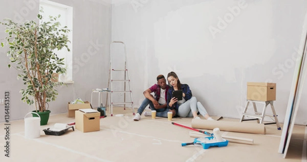 Fototapeta A loving couple relaxes on the floor of their newly renovated apartment, drinking take-out coffee and scrolling through a tablet for furniture and decoration ideas.