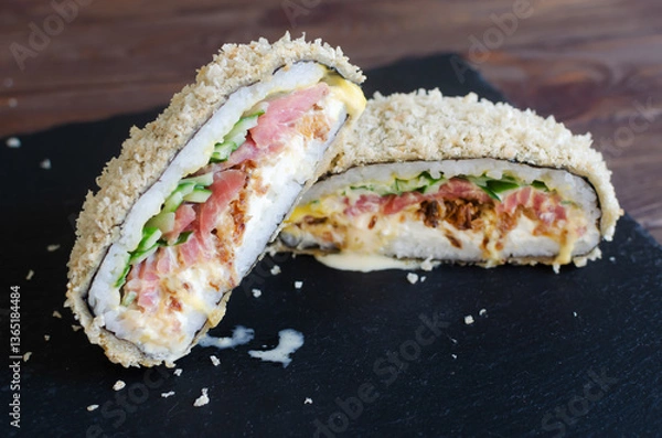 Fototapeta Sushi burger with tuna, cucumber, onion, cheese and rice in nori with crispy crust on a black plate on a wooden table. Creative dish. Hybrid food concept. Horizontal orientation. Selective focus.