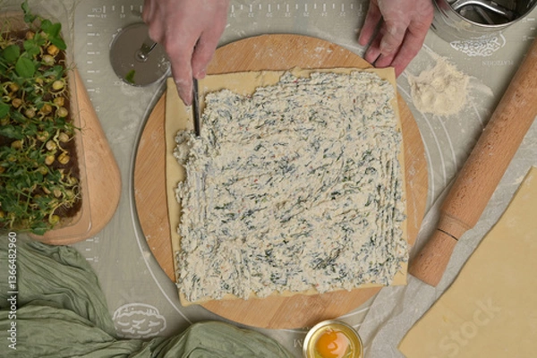 Obraz Woman's hand with fork putting cottage cheese with parsley and dill greens on dough. Rolling pin, cutter, egg, green towel and microgreens sprouts on kitchen table. Top view. Home cooking baking puffs
