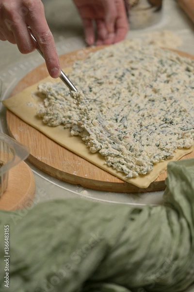Obraz Woman's hand with fork putting cottage cheese with parsley and dill greens on dough. Green towel, wooden board on kitchen table. Close up. Home cooking baking puffs