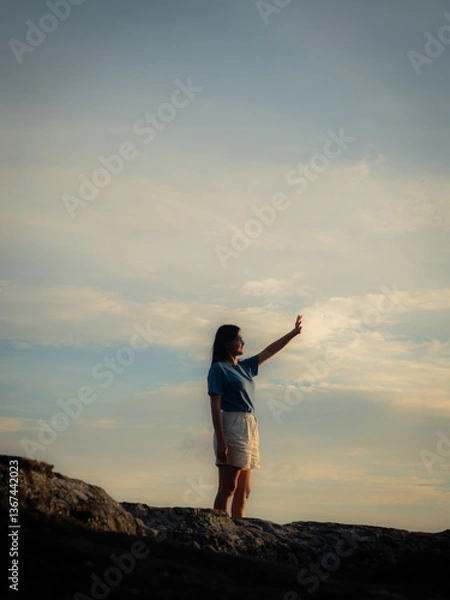 Fototapeta Woman stands watching the sunset on the edge of a cliff in a large sky filled with thin clouds and warm golden light