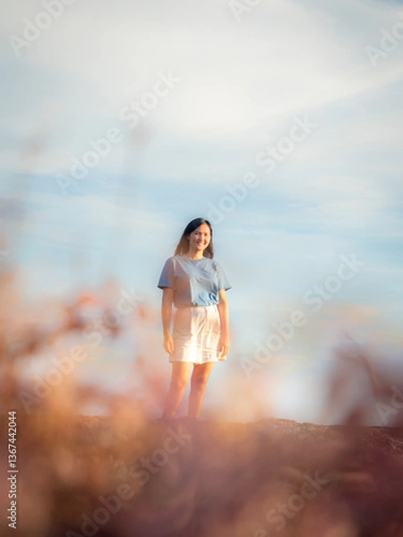 Fototapeta Woman stands watching the sunset on the edge of a cliff in a large sky filled with thin clouds and warm golden light