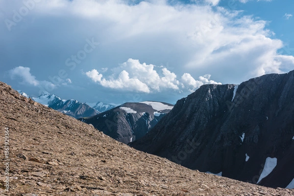 Fototapeta Dramatic vast view from sunlit high stony pass to big rocky top with snowfield, large snowy mountain range silhouette with pointy peaks far away under clouds in blue sky. Sheer crags and peaked tops.