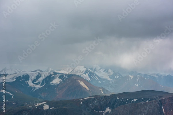 Fototapeta Dark atmospheric landscape with mountain silhouettes and large snow-capped peaked top in rainy low clouds. Dramatic alpine view to high mountains in gray cloudy sky. Big snowy mountain range in rain.
