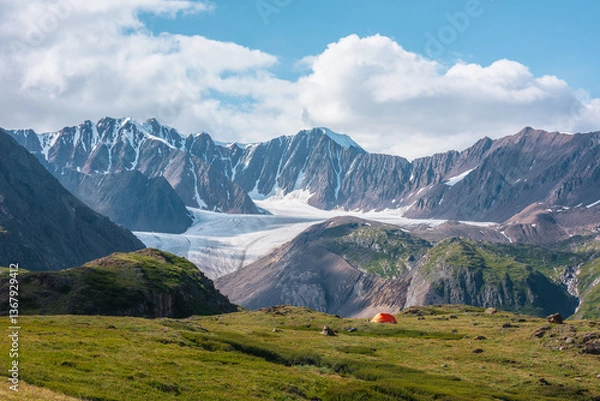 Fototapeta Colorful landscape with orange tent in alpine valley with view to big glacier tongue and large snow-capped mountain range under clouds in blue sky. Green hills and rocks against ice and sheer crags.