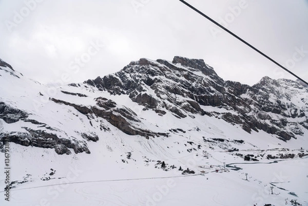 Fototapeta A photo of a snow-covered mountain peak in winter, with a cable car line visible on the right side of the image.