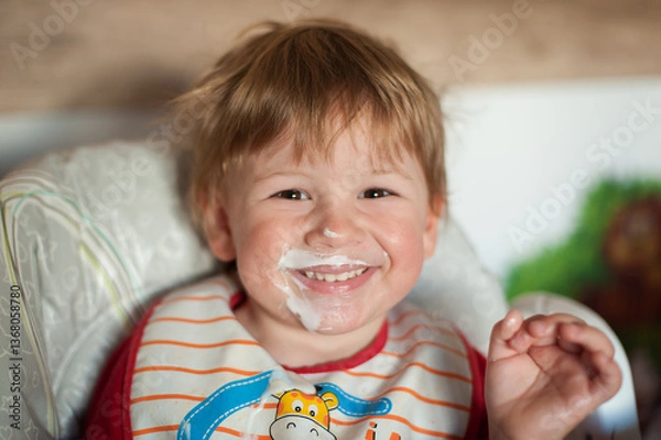 Fototapeta A toddler sits happily in a high chair, wearing a bib and smiling with yogurt on his face. The warm morning light highlights his cheerful expression in a cozy kitchen atmosphere