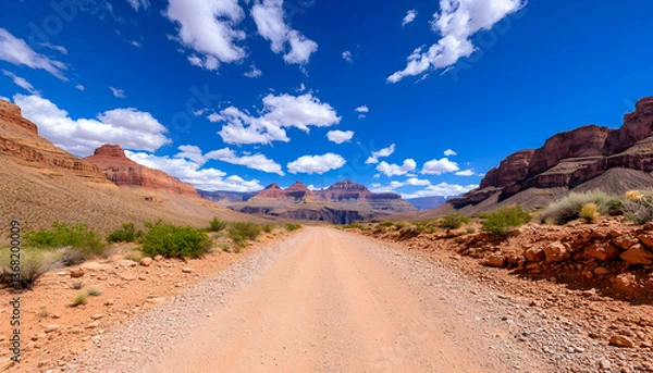 Fototapeta Scenic desert dirt road leading into a vast canyon under a vibrant blue sky with fluffy clouds