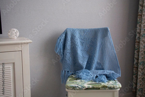 Fototapeta A blue shawl knitted using the fillet knitting technique hangs on the back of a chair against a gray wall.