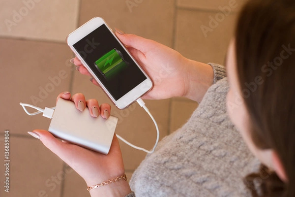 Fototapeta Girl hold in hands smart phone and power bank. Charge smart phone from power bank. Use modern gadget.