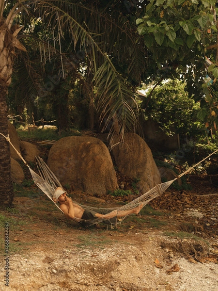 Fototapeta A man in a Hammock lies and smokes legal Marijuana in Thailand under the palm trees.