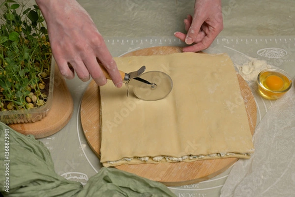 Obraz Square sheet of dough cut into strips with woman's hands on wooden board. Cutter, egg, green towel, microgreens sprouts on kitchen table. Home cooking baking puffs