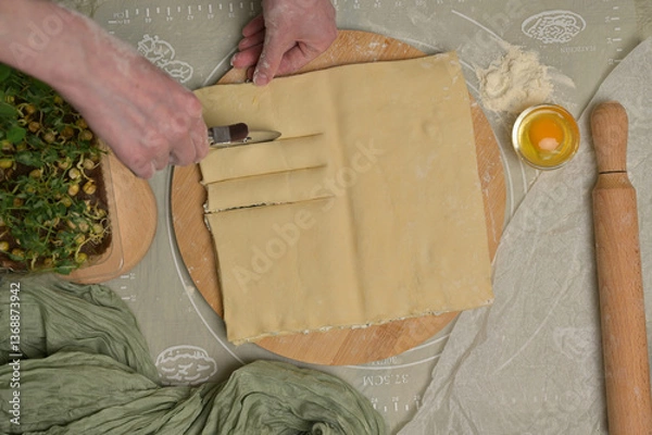 Obraz Top view square sheet of dough cut into strips with woman's hands on wooden board. Cutter, egg, green towel, microgreens sprouts on kitchen table. Home cooking baking puffs