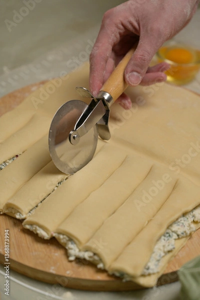 Obraz Close up square sheet of dough cut into strips with woman's hands on wooden board. Cutter, egg on kitchen table. Home cooking baking puffs