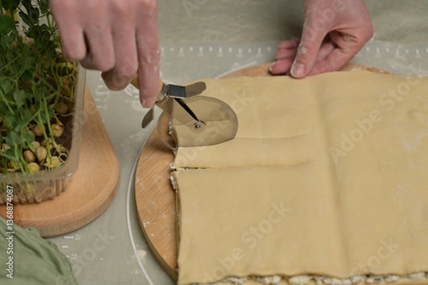 Obraz Close up square sheet of dough cut into strips with woman's hands on wooden board. Cutter, green towel, microgreens sprouts on kitchen table. Home cooking baking puffs