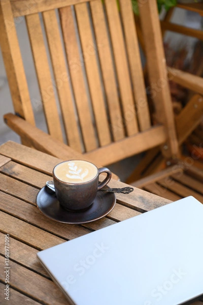 Fototapeta A modern workspace with a laptop and cappuccino on a wooden table in an outdoor setting. Warm natural tones and soft lighting create a productive yet relaxing atmosphere for remote work.