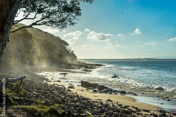 Fototapeta Tropical Surfer Beach. Idyllic sunset at famous Tea Tree Bay, Noosa, Australia. Sun shining through spray mist in the eucalyptus trees. Tropical stone pebbles in the foreground. Two surfers ret