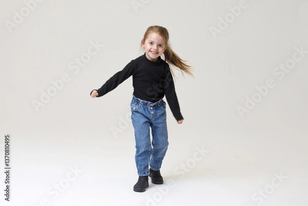 Fototapeta Happy little girl dancing with joy. style - Black sweater, blue jeans, and boots, smiling and moving energetically against a neutral background.