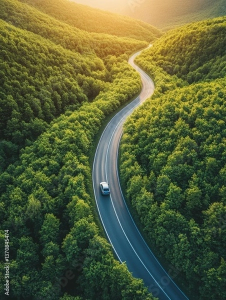 Fototapeta Aerial view of a car driving on a winding road through lush green forest at sunset in a scenic landscape