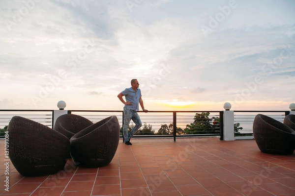 Fototapeta handsome guy standing on balcony in luxury hotels in tropics at sunset and drinking tea