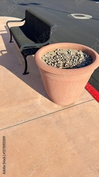 Obraz empty metal bench beside terracotta pot in quiet parking lot scene with painted lines sunlit pavement concrete surface peaceful afternoon minimal urban setting with subtle earthy tones